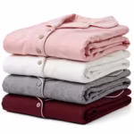 Folded stack of four ladies pyjama sets in soft pink, white, grey marl and burgundy cotton jersey, each folded as a pair with the button-front top over its pants, white piping and matte buttons showing