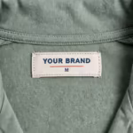 Close-up of the inside back neck of the ladies sage green pyjama top showing the cream woven YOUR BRAND label with navy lettering, a red rule line and the size M mark, stitched into the inside back neck below the collar band on the reverse side of the sage cotton single jersey