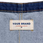 Close-up of the inside back neck of the mid-indigo denim bib overalls showing the cream woven YOUR BRAND label with navy lettering, a red rule line and the size M mark, stitched into the inside of the bib just below its top hem seam