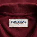 Close-up of the inside back neck of the corporate polo shirt showing the cream woven YOUR BRAND label with navy lettering, a red rule line and the size M mark, stitched into the neck binding