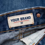 Close-up of the inside back waistband of the denim pants showing the cream woven YOUR BRAND label with navy lettering, a red rule line and the size 32 mark, stitched into the waistband