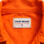 Close-up of the inside back neck of the safety orange flame resistant coverall, showing the cream woven YOUR BRAND label with navy lettering, red rule line and size M mark, sewn in below the collar band stitching