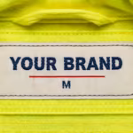 Close-up of the inside back neck of the fluorescent yellow high visibility jacket, showing the cream woven YOUR BRAND label with navy lettering, a red rule line and the size M mark, stitched into the neck seam