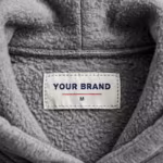 Close-up of the inside back neck of the grey marl brushed fleece pullover hoodie showing the cream woven YOUR BRAND label with navy lettering, a red rule line and the size M mark, stitched into the inside back neck seam below the hood