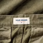 Close-up of the inside back waistband of the olive hunting trousers, showing the cream woven YOUR BRAND label with navy lettering, a red rule line and the size 32 mark, stitched into the waistband