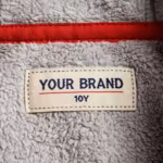 Close-up of the inside back neck of the red kids softshell jacket showing the cream woven YOUR BRAND label with navy lettering, a red rule line and the size 10Y mark, stitched into the grey brushed fleece backing at the inside neck seam