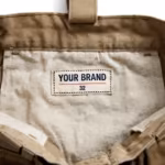 Close-up of the inside back waistband of the khaki kneepad work trousers showing the cream woven YOUR BRAND label with navy lettering, a red rule line and the size 32 mark, stitched into the waistband