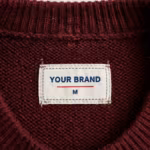 Close-up of the inside back neck of the burgundy knitted crew-neck pullover showing the cream woven YOUR BRAND label with navy lettering, a red rule line and the size M mark, stitched into the reverse side of the knit just below the ribbed crew neckband