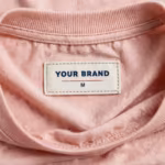 Close-up of the inside back neck of the soft pink maternity t-shirt showing the cream woven YOUR BRAND label with navy lettering, a red rule line and the size M mark, stitched into the soft pink jersey just below the ribbed crew neck band