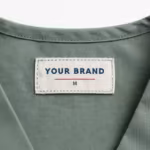 Close-up of the inside back neck of the sage green unisex medical scrub top showing the cream woven YOUR BRAND label with navy lettering, a red rule line and the size M mark, stitched into the inside back neckband of the sage poly-cotton twill