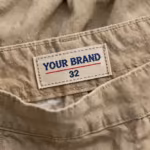 Close-up of the inside back waistband of the khaki canvas work shorts showing the cream woven YOUR BRAND label with navy lettering, a red rule line and the size 32 mark, stitched into the waistband