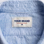 Close-up of the inside back neck of the casual shirt showing the cream woven YOUR BRAND label with navy lettering, a red rule line and the size M mark, stitched into the collar band