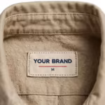 Close-up of the inside back neck of the men's stone cotton twill utility jacket showing the cream woven YOUR BRAND label with navy lettering, a red rule line and the size M mark, stitched into the inside collar band