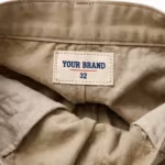 Close-up of the inside back waistband of the men's khaki cotton twill shorts showing the cream woven YOUR BRAND label with navy lettering, a red rule line and the size 32 mark, stitched into the waistband
