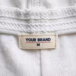 Close-up of the inside back waistband of the white ladies cotton classic brief showing the cream woven YOUR BRAND label with navy lettering, a red rule line and the size M mark, stitched into the waistband
