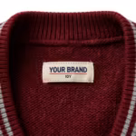 Close-up of the inside back neck of the burgundy knitted school sweater showing the cream woven YOUR BRAND label with navy lettering, a red rule line and the size 10Y mark, stitched flat into the centre of the inside back neck just below the ribbed neck band on the reverse purl side of the burgundy knit