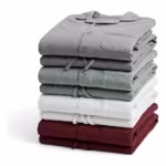Folded stack of the same men's two-piece pyjama sets in grey marl, sage green, white and burgundy cotton jersey, each folded top showing the notched collar, button placket and chest pocket
