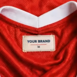 Close-up of the inside back neck of the red sports jersey showing the cream woven YOUR BRAND label with navy lettering, a red rule line and the size M mark, stitched into the red eyelet mesh below the white ribbed V-neck collar band
