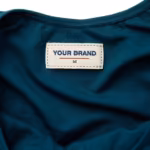 Close-up of the inside back of the deep teal ladies one-piece swimsuit showing the cream woven YOUR BRAND label with navy lettering, a red rule line and the size M mark, stitched inside the back below the neckline