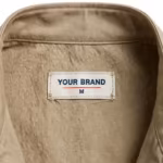Close-up of the inside back neck of the khaki cotton twill work vest showing the cream woven YOUR BRAND label with navy lettering, a red rule line and the size M mark, stitched into the inside back neck below the stand collar seam