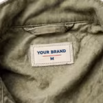 Close-up of the inside back neck of the olive cotton twill work jacket showing the cream woven YOUR BRAND label with navy lettering, a red rule line and the size M mark, stitched into the centre of the inside back neck just below the stand collar band